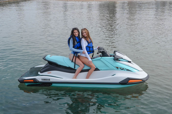 Two friends wearing blue life jackets smiling while seated on a turquoise-and-white jet ski on calm coastal waters near a sandy shoreline