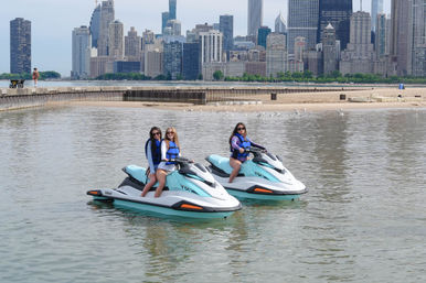 Three people in life jackets on teal-and-white jet skis near a sandy lakefront beach with the Chicago skyline in the background.