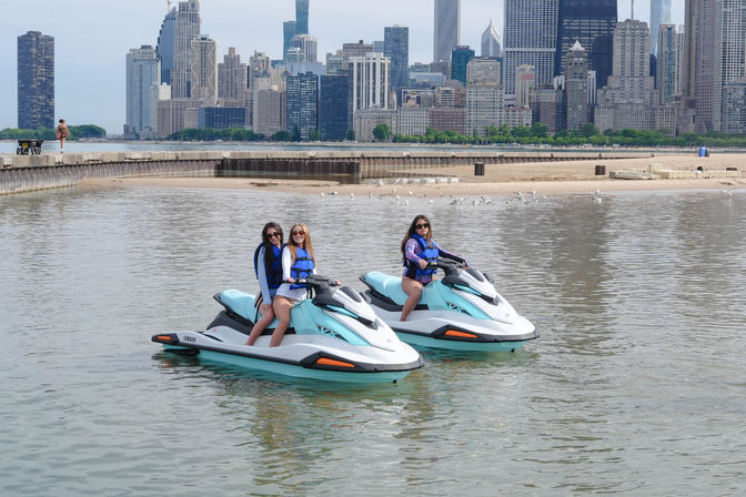Three people in life jackets on teal-and-white jet skis near a sandy lakefront beach with the Chicago skyline in the background.