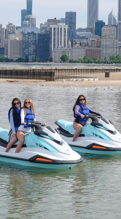 Three people in life jackets on teal-and-white jet skis near a sandy lakefront beach with the Chicago skyline in the background.