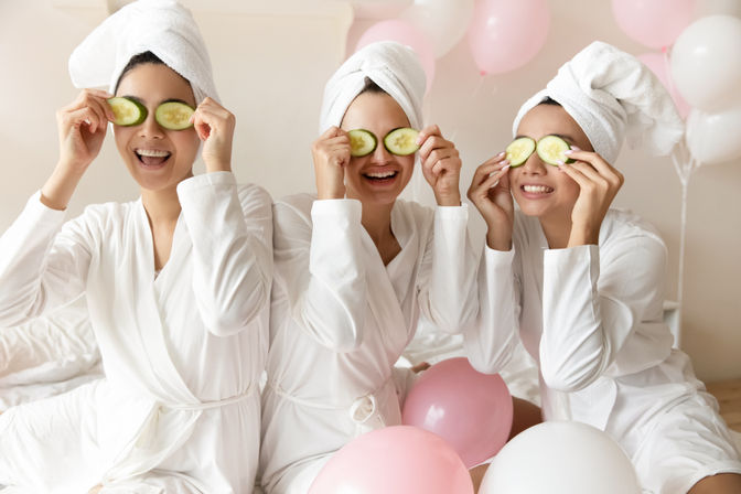 Three friends in white robes and towel turbans laughing and holding cucumber slices over their eyes during a fun at-home spa day with pink and white balloons.