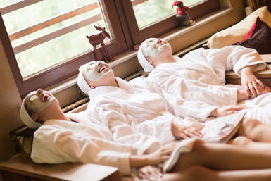 Three friends in white robes with facial masks and cucumber eye slices laughing and lounging on a sunlit spa lounge by a window