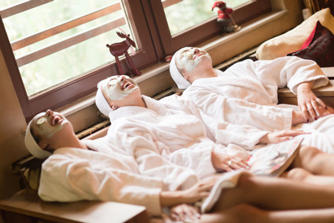 Three friends in white robes with facial masks and cucumber eye slices laughing and lounging on a sunlit spa lounge by a window