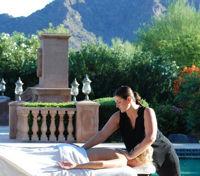 Poolside outdoor massage: a therapist giving a back massage to a client on a table beside a turquoise pool, framed by ornamental stone balustrade, lush gardens and desert mountains in the background.