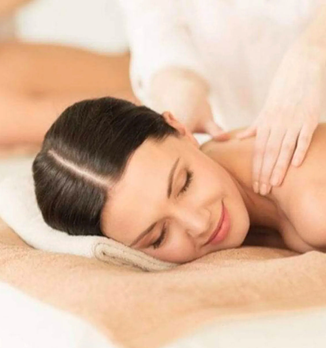 Relaxed woman lying face down on a towel with eyes closed, receiving a shoulder massage at a spa — peaceful relaxation and wellness treatment.
