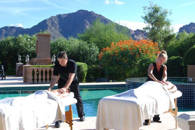 Poolside spa scene with two therapists giving outdoor massages on tables beside a turquoise pool, orange flowering shrubs and lush greenery, and rugged desert mountains under a sunny blue sky.