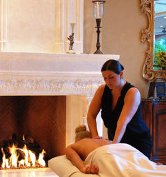 Client receiving a back massage from a therapist in a luxurious indoor spa suite beside a roaring fireplace, ornate stone mantel and warm ambient lighting