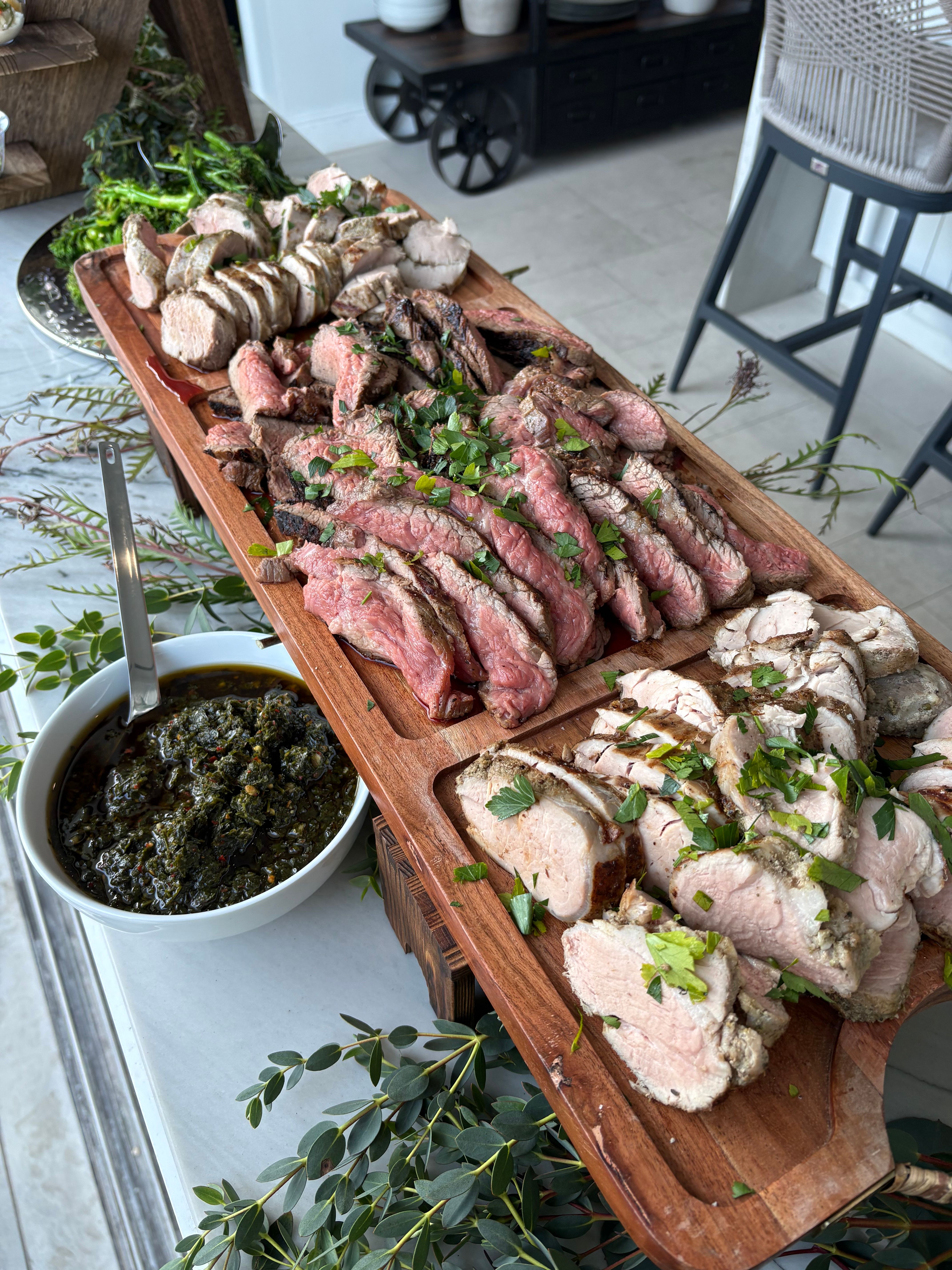 Sliced medium-rare steak and roasted pork arranged on a wooden serving board, garnished with chopped parsley and served with chimichurri in white bowls on a bright patio table.