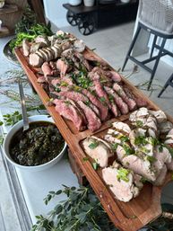Sliced medium-rare steak and roasted pork arranged on a wooden serving board, garnished with chopped parsley and served with chimichurri in white bowls on a bright patio table.