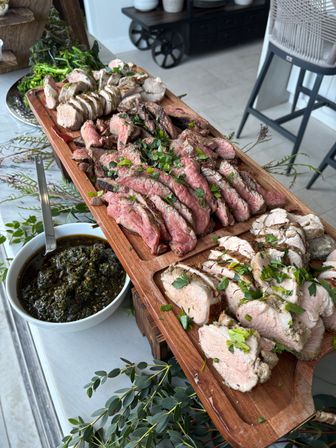 Sliced medium-rare steak and roasted pork arranged on a wooden serving board, garnished with chopped parsley and served with chimichurri in white bowls on a bright patio table.