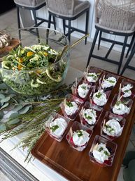 Glass bowl of fresh arugula salad with citrus and fennel and a gold serving spoon beside a wooden tray of individual berry parfaits topped with whipped cream and mint, arranged on a leafy-decorated patio counter with woven bar stools in the background