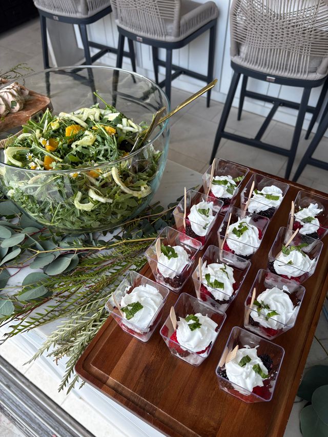 Glass bowl of fresh arugula salad with citrus and fennel and a gold serving spoon beside a wooden tray of individual berry parfaits topped with whipped cream and mint, arranged on a leafy-decorated patio counter with woven bar stools in the background