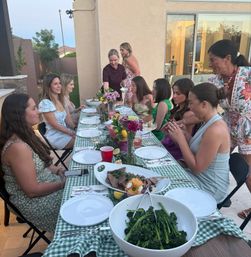 Outdoor backyard patio dinner party at dusk — women and girls seated at a long green-check table with floral centerpieces, bowls of broccolini and sliced steak, plates and drinks near a sliding glass door in a suburban home.