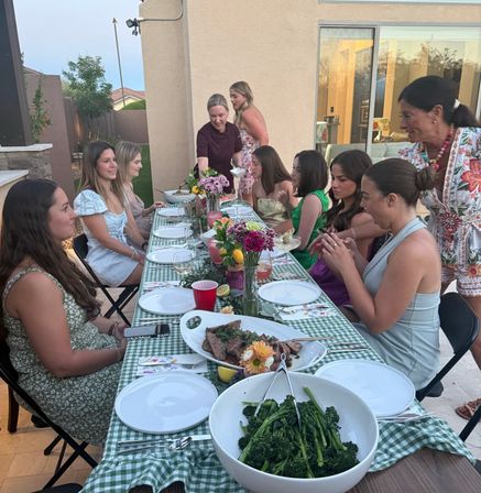 Outdoor backyard patio dinner party at dusk — women and girls seated at a long green-check table with floral centerpieces, bowls of broccolini and sliced steak, plates and drinks near a sliding glass door in a suburban home.