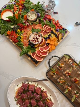 Colorful grazing board and appetizer spread: wooden charcuterie platter with sliced grapefruit, strawberries, cantaloupe, peaches, blueberries, cherry tomatoes, radishes, snap peas, edible flowers and two creamy dips, served beside shrimp cocktail shooters and prosciutto-wrapped bites on a white serving dish.