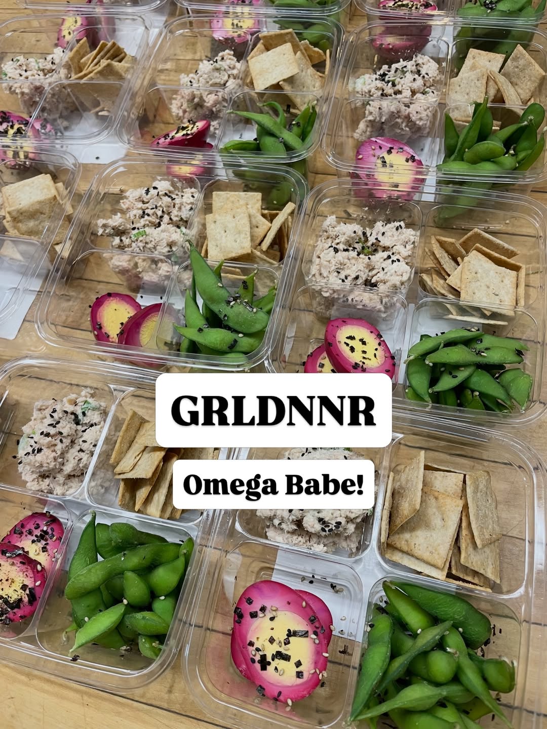 Clear meal-prep boxes on a wooden table with tuna salad topped with sesame seeds, bright green edamame, crispy crackers and vibrant pink pickled eggs — healthy grab-and-go snack boxes.