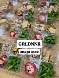 Clear meal-prep boxes on a wooden table with tuna salad topped with sesame seeds, bright green edamame, crispy crackers and vibrant pink pickled eggs — healthy grab-and-go snack boxes.