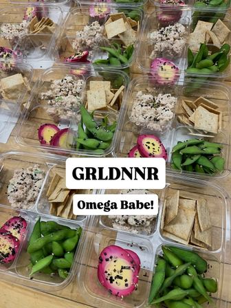 Clear meal-prep boxes on a wooden table with tuna salad topped with sesame seeds, bright green edamame, crispy crackers and vibrant pink pickled eggs — healthy grab-and-go snack boxes.