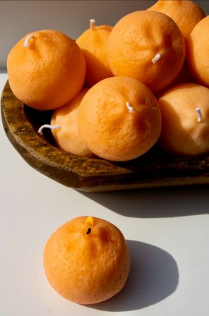 Orange spherical candles used as tabletop decor — several nestled in a rustic wooden bowl and one lit on a white surface casting a warm glow and soft shadow.
