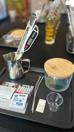 DIY candle-making station on black tray: stainless pouring pitcher with digital thermometer, packaged bamboo stirrers, pre-tabbed wicks, glass jar with bamboo lid, small mixing cup, markers and alcohol pad.