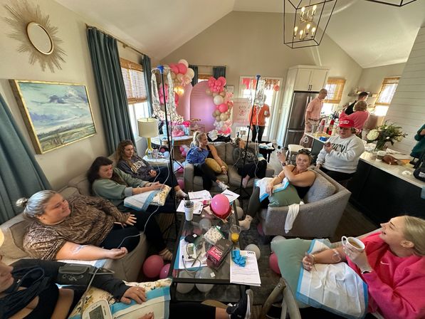 At-home group IV hydration party in a cozy living room — women lounging on sofas receiving IV drips amid pink and gold balloon arch and celebratory decorations.