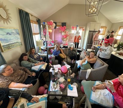 At-home group IV hydration party in a cozy living room — women lounging on sofas receiving IV drips amid pink and gold balloon arch and celebratory decorations.
