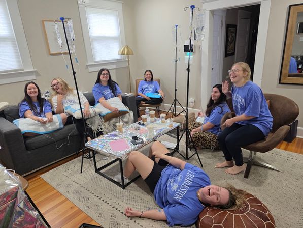 Seven women in matching blue 'Charleston' shirts laughing in a cozy living room, gathered around a coffee table and enjoying a playful at-home IV hydration drip session with IV bags on stands.