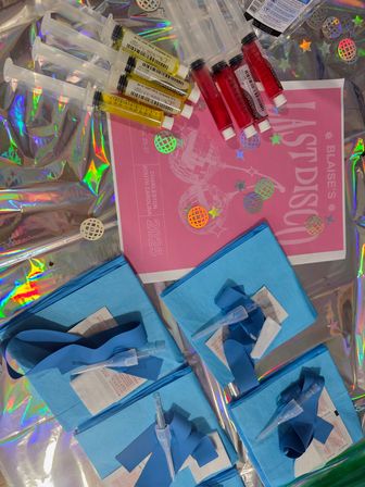 Overhead view of colorful syringes filled with red and yellow liquids, blue sterile packs with tourniquets and needle caps, and a pink disco-themed flyer arranged on holographic foil.
