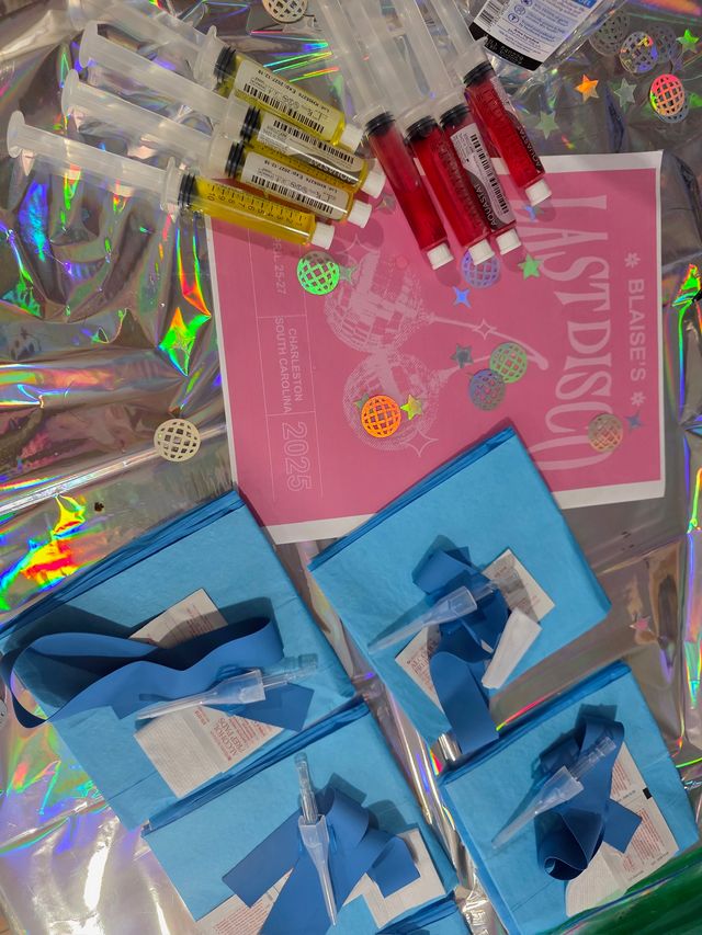 Overhead view of colorful syringes filled with red and yellow liquids, blue sterile packs with tourniquets and needle caps, and a pink disco-themed flyer arranged on holographic foil.