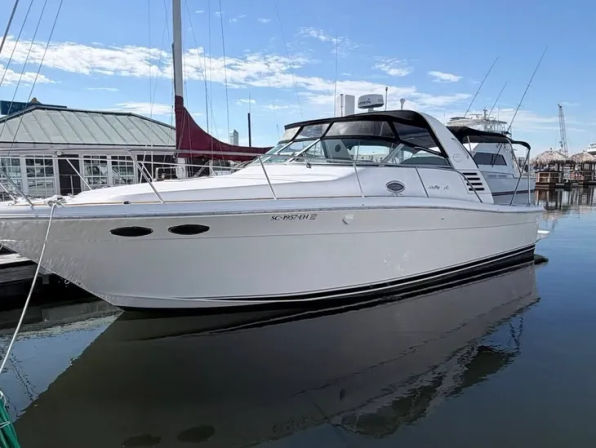 White luxury cabin-cruiser yacht docked at a coastal marina, sleek black canvas hardtop and windows reflecting in calm water under a sunny blue sky
