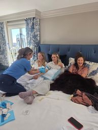 Four women lounging on a king bed in a bright hotel-style bedroom as a medical professional administers IV therapy; friends laugh and sip drinks by blue patterned curtains
