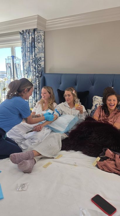 Four women lounging on a king bed in a bright hotel-style bedroom as a medical professional administers IV therapy; friends laugh and sip drinks by blue patterned curtains