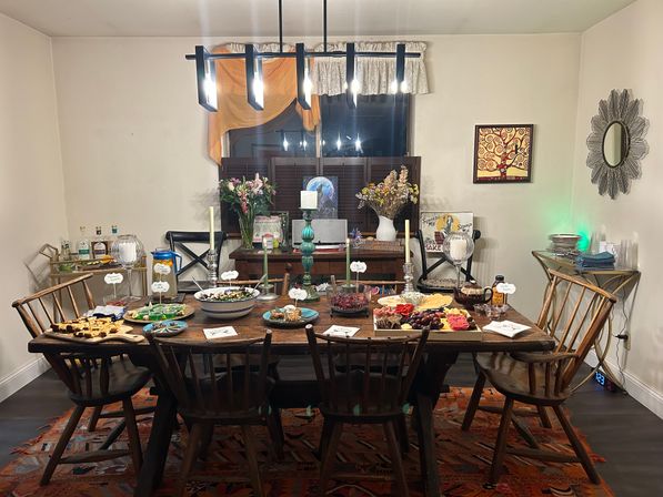 Cozy dining room with a large wooden farmhouse table laid out as a party-ready buffet — charcuterie, salads, bowls and candles under a modern black chandelier, surrounded by wooden chairs and wall decor.
