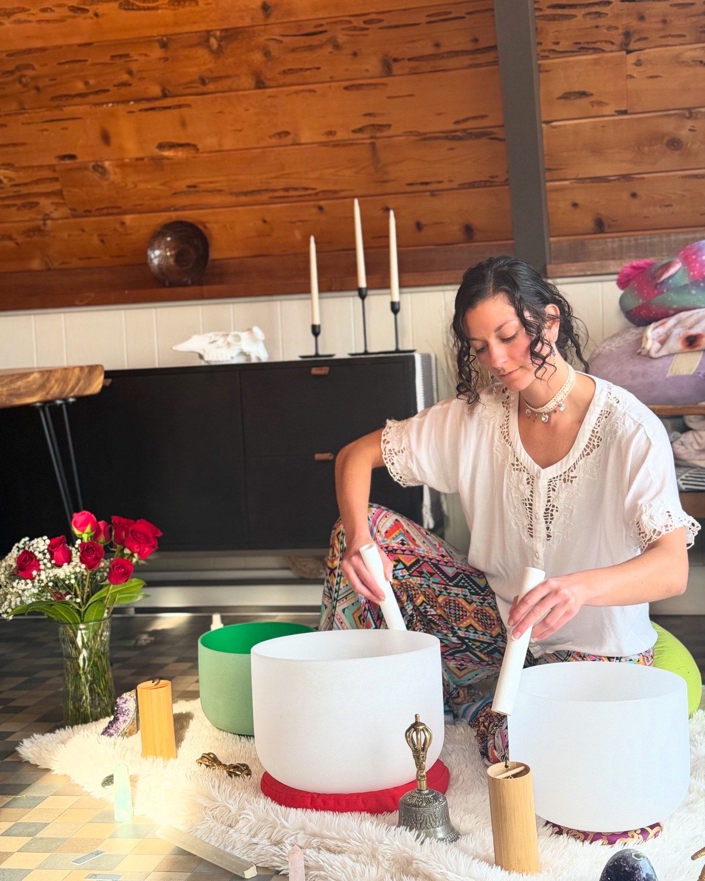 Woman playing crystal singing bowls in a cozy wood-paneled studio, sound healing setup with candles, meditation tools and a vase of red roses, wearing a white blouse and colorful patterned pants.