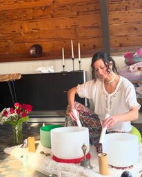 Woman playing crystal singing bowls in a cozy wood-paneled studio, sound healing setup with candles, meditation tools and a vase of red roses, wearing a white blouse and colorful patterned pants.