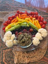 Vibrant snack platter of cherry tomatoes, baby carrots, yellow bell pepper strips, celery, cauliflower florets, blueberries and grapes surrounding a glass bowl of cannabis buds on a patterned tablecloth.