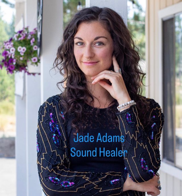 Portrait of a smiling woman with curly dark hair and a septum ring on a sunny porch beside hanging purple petunias, wearing a beaded boho jacket and bracelets.