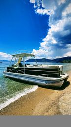 Sleek pontoon boat beached on a sandy alpine lake shore with sparkling turquoise water, forested hills on the horizon and towering cumulus clouds against a vivid blue sky