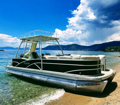 Sleek pontoon boat beached on a sandy alpine lake shore with sparkling turquoise water, forested hills on the horizon and towering cumulus clouds against a vivid blue sky