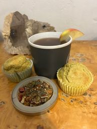 Cozy tea scene on a rustic wood table: a black paper cup of hot tea with an apple slice garnish, two greenish muffins in paper liners, a tin lid of loose herbal tea, and a smoky crystal display.