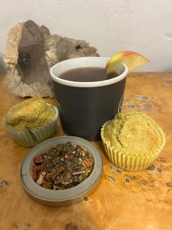 Cozy tea scene on a rustic wood table: a black paper cup of hot tea with an apple slice garnish, two greenish muffins in paper liners, a tin lid of loose herbal tea, and a smoky crystal display.