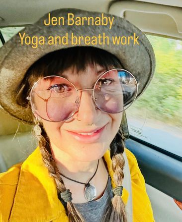 Close-up selfie in a car of a smiling person wearing a gray felt hat, round pink-tinted glasses, braided pigtails, a nose ring and silver pendant, dressed in a yellow jacket; yellow text overlay promoting yoga and breath work.