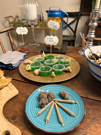 Home gathering spread on a wooden table: blue plate of cannabis pre-rolls and nugs fanned out, gold platter of bright green jello shots garnished with lime and ginger, citrus-infused water pitcher and salad bowl nearby.