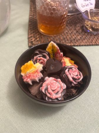 Autumn-themed chocolates shaped like roses and colorful leaves in a black bowl on a table, with a honey-colored jar on a woven placemat in the background