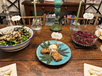 Rustic wooden table with a family-style dinner spread: mixed green salad in a blue-rimmed bowl labeled Lettuce, crystal bowl of roasted beets labeled The Beets, turquoise plate of crumble with vanilla ice cream and decorative leaves, and green taper candles.