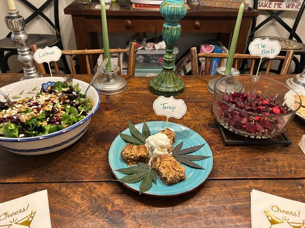 Rustic wooden table with a family-style dinner spread: mixed green salad in a blue-rimmed bowl labeled Lettuce, crystal bowl of roasted beets labeled The Beets, turquoise plate of crumble with vanilla ice cream and decorative leaves, and green taper candles.