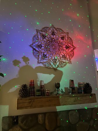 Wooden mandala wall art above a rustic mantel shelf with candles, pinecones and small decorative figures, bathed in colorful light and green laser star dots from a projector