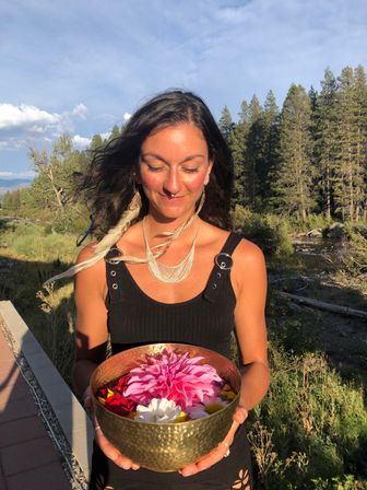 Smiling woman with feather earrings holding a hammered brass bowl of bright flowers in a sunlit mountain river valley with pine trees