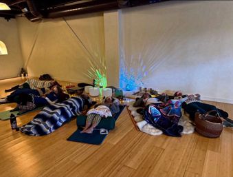 Group sound bath meditation in a yoga studio — people lying on mats and bolsters under blankets on a warm wooden floor, surrounded by crystal singing bowls, soft string lights and colorful wall light projections.