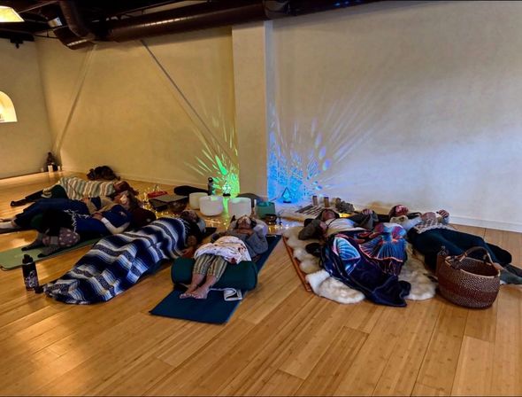 Group sound bath meditation in a yoga studio — people lying on mats and bolsters under blankets on a warm wooden floor, surrounded by crystal singing bowls, soft string lights and colorful wall light projections.